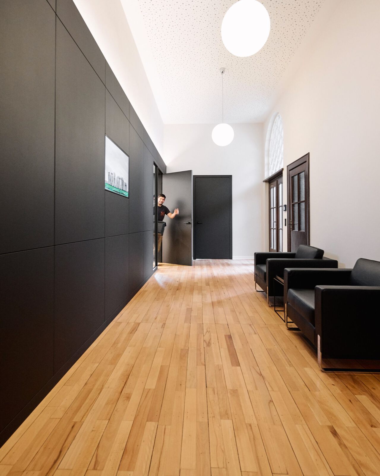 Moderner B&uuml;roflur mit dunklen Wandpaneelen, Holzboden und schwarzen Sitzm&ouml;beln rechts.