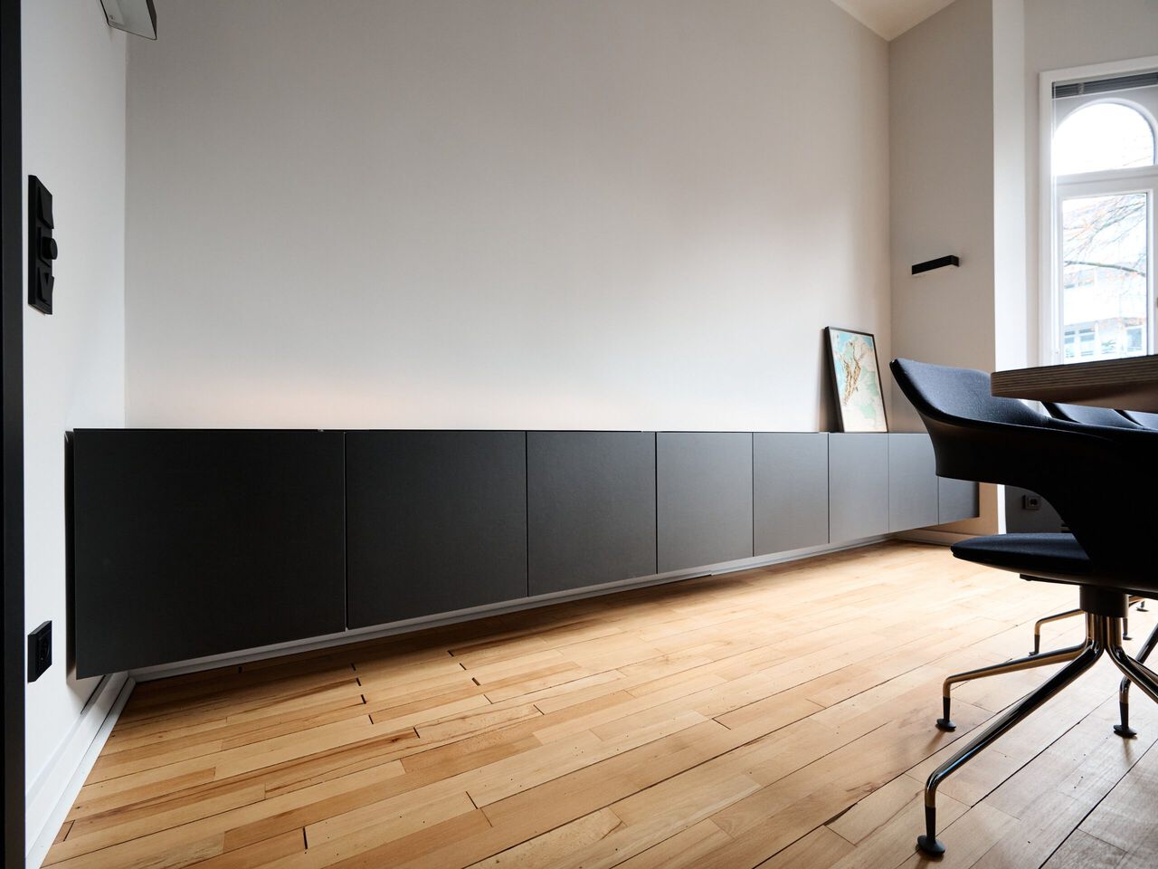 Modernes B&uuml;ro mit schwarzem Sideboard, Holzboden und gro&szlig;em Fenster.