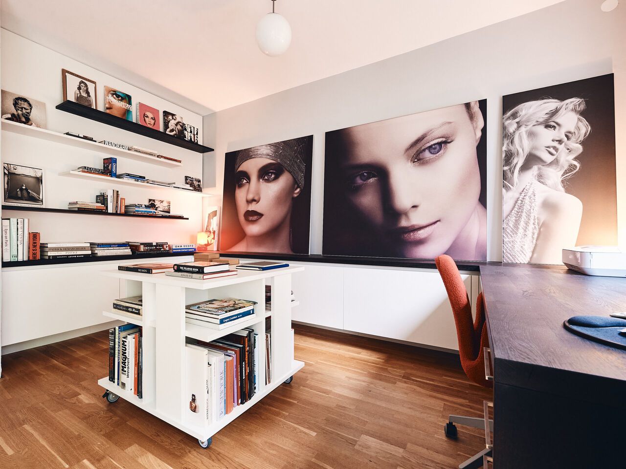 Modernes B&uuml;ro mit wei&szlig;en Wandregalen, gro&szlig;formatigen Portr&auml;ts und zentralem wei&szlig;em B&uuml;cherregal auf Holzboden.