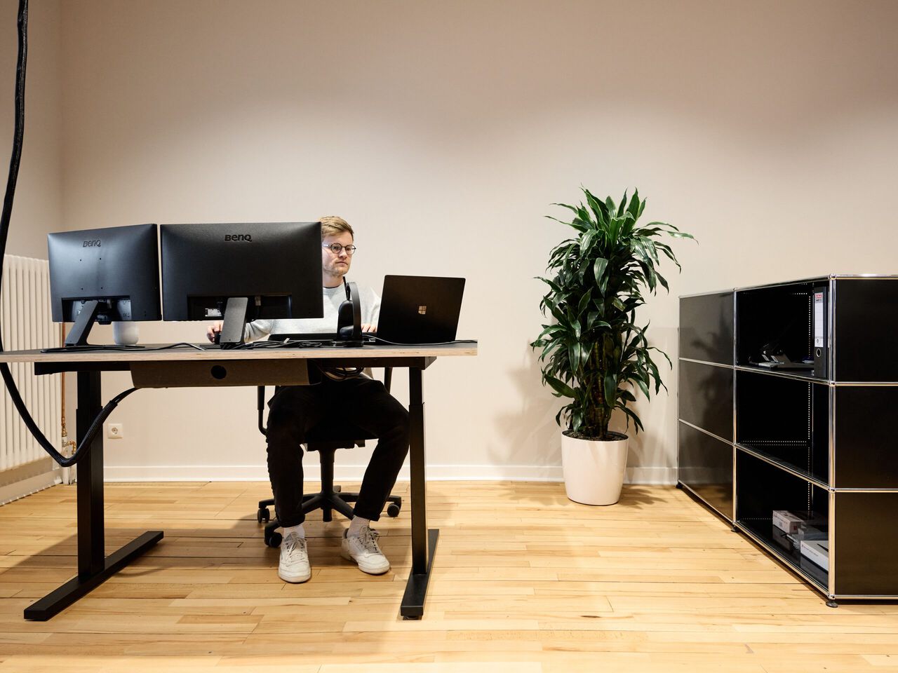Person sitzt an schwarzem Schreibtisch mit Monitoren und Laptop in modernem B&uuml;ro mit Holzboden und Pflanze.