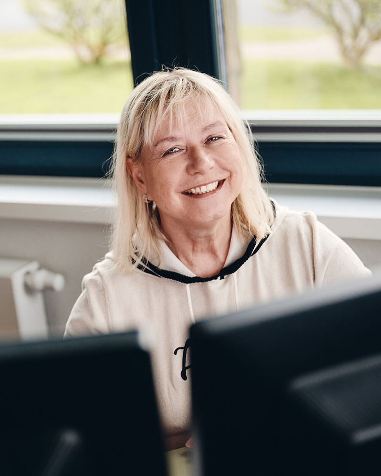 Eine Frau mit blonden Haaren sitzt l&auml;chelnd vor einem Computer am Fenster.