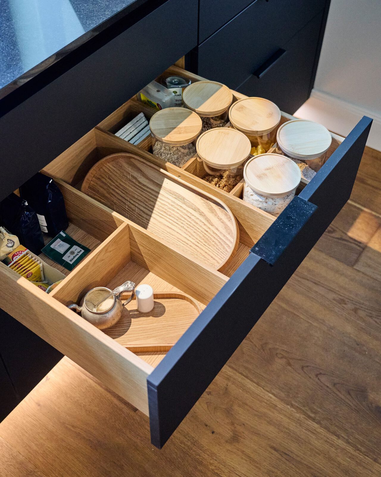 Offene Schublade einer schwarzen Küchenkommode mit Holzorganizern für Behälter, Bretter und Küchenutensilien.
