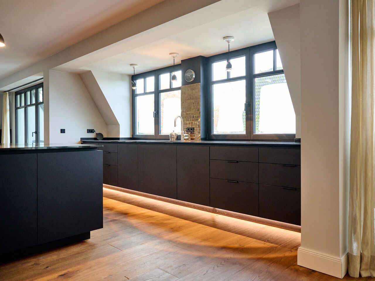 Moderne schwarze Küchenzeile mit LED-Bodenbeleuchtung, großen Fenstern und Holzparkett.