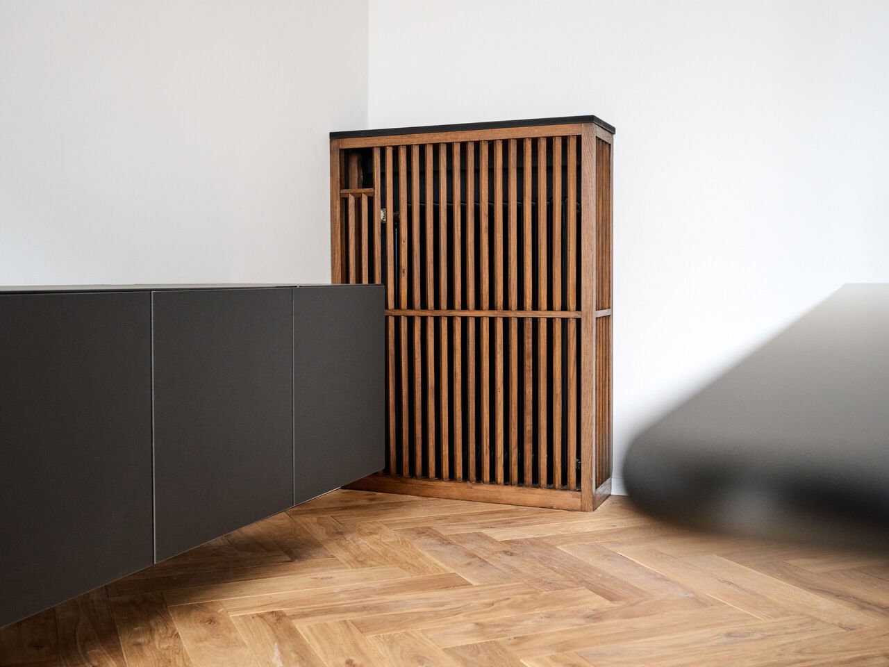 Holzschrank mit vertikalen Lamellen in einem minimalistischen Raum mit Parkettboden und dunkler Wandverkleidung.