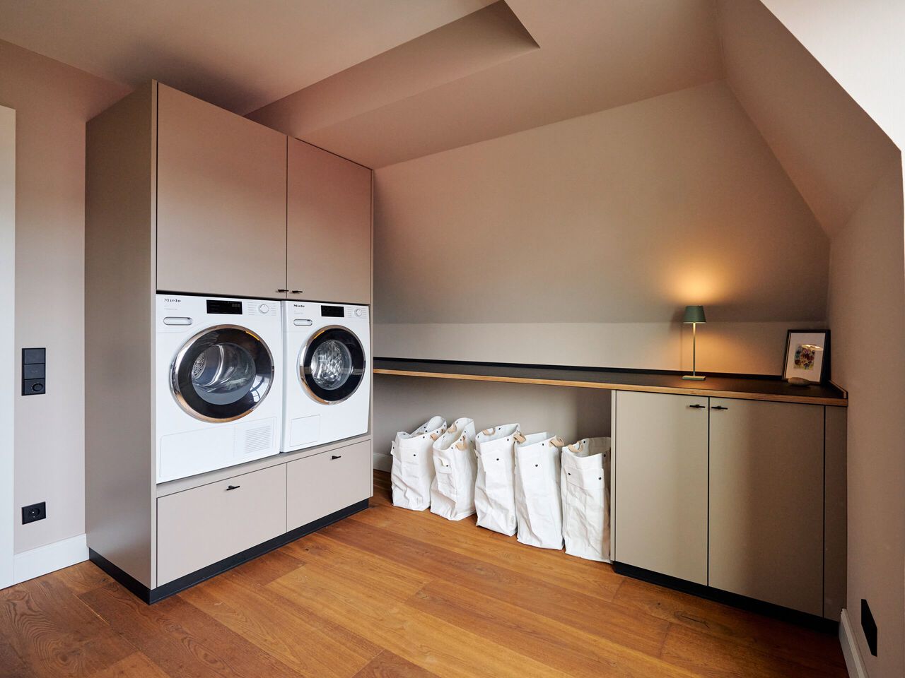 Moderne Waschküche mit zwei weißen Waschmaschinen auf erhöhten Sockeln, Holzboden und Aufbewahrungsschränken für Wäsche.