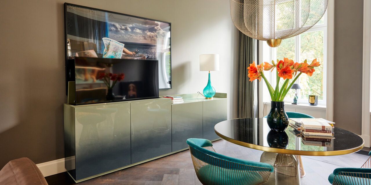 Modernes Wohnzimmer mit grauem Sideboard, Fernseher, rundem Tisch und Blumenvase am Fenster.