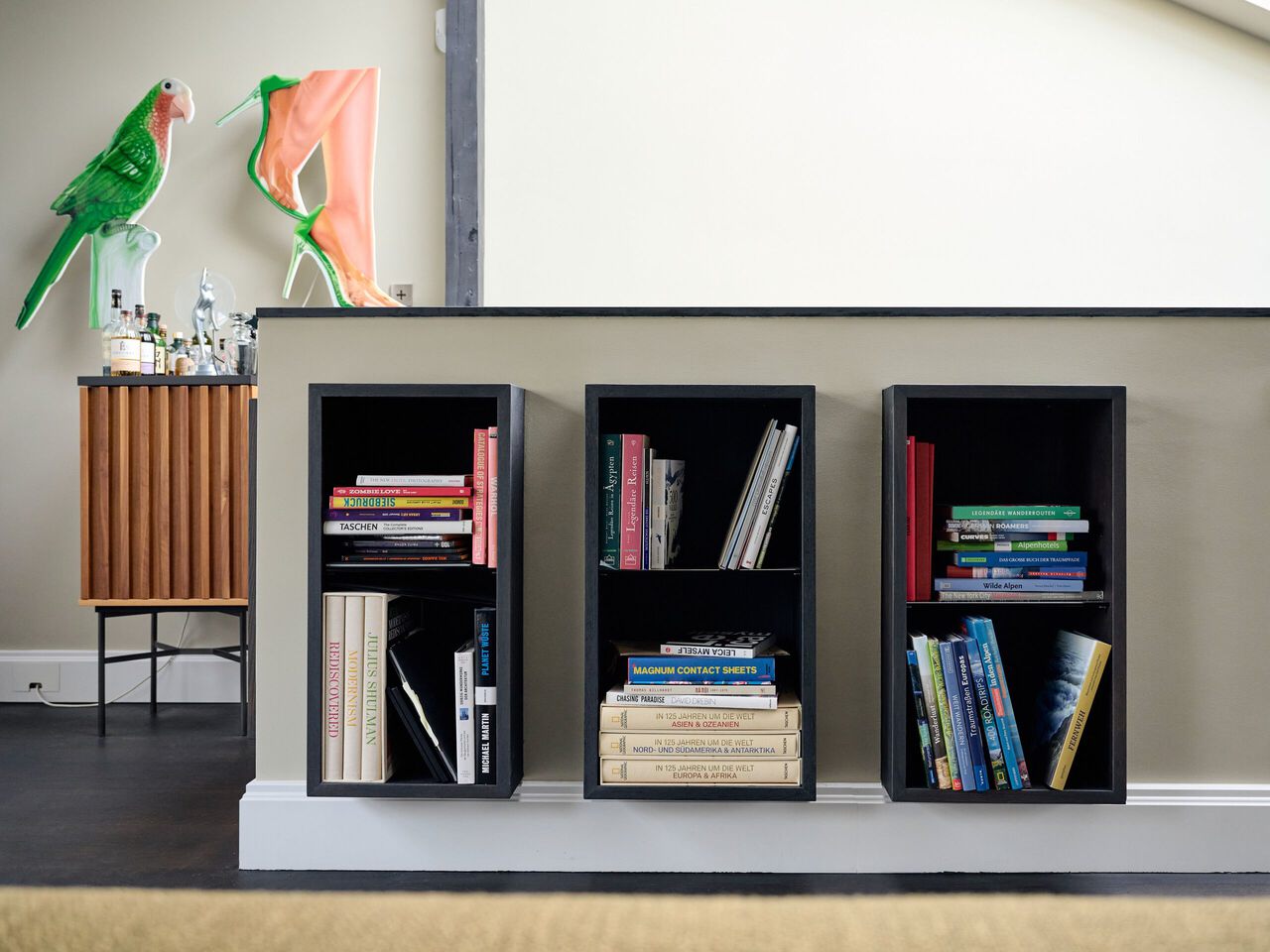 Drei schwarze Bücherregale mit Büchern, daneben ein Holzschrank mit grünem Papagei und Kunstwerk an der Wand.