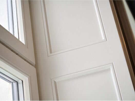Weiße Innentür mit rechteckigen Kassetten und angrenzenden Fensterreihen in heller Raumecke.