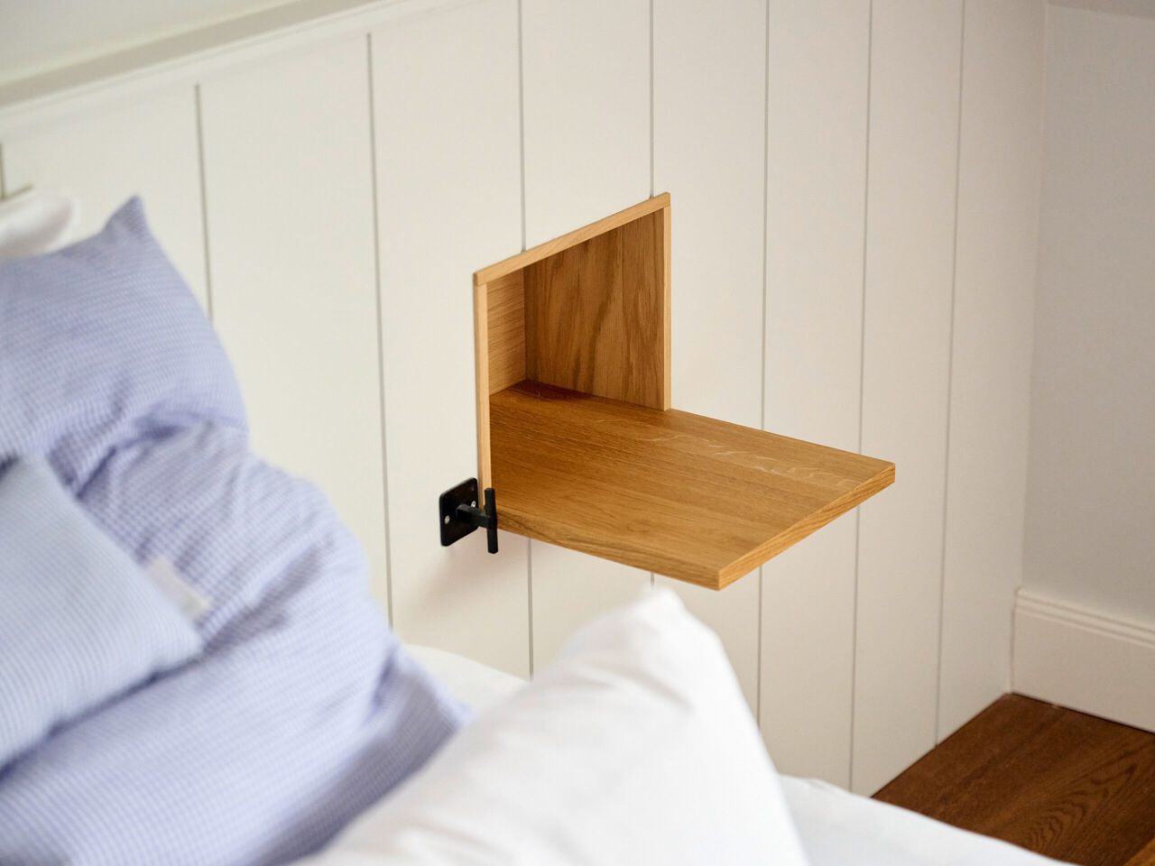 Wandmontierter Holzklapptisch neben einem Bett mit blauem Kissen und wei&szlig;er Bettw&auml;sche.