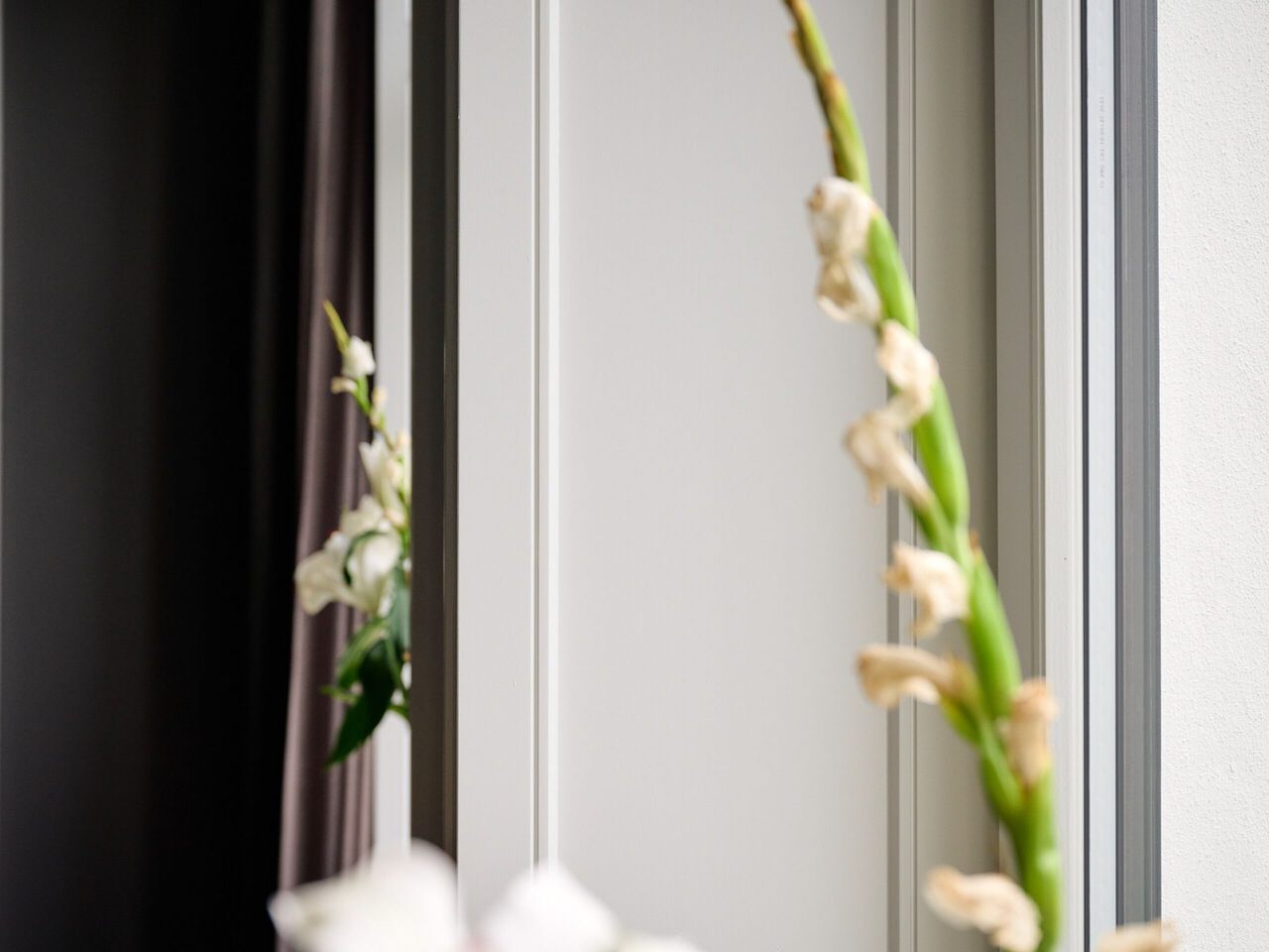 Wei&szlig;e Bl&uuml;ten an gr&uuml;nen Stielen in einer modernen Wohnraumecke neben Fenster und dunklem Vorhang.