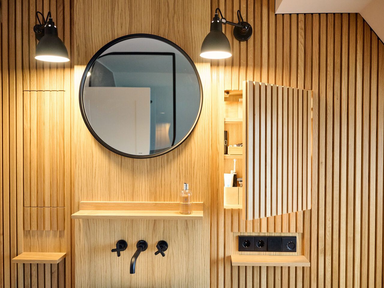 Modernes Badezimmer mit Holzverkleidung, rundem Spiegel und schwarzen Wandarmaturen.