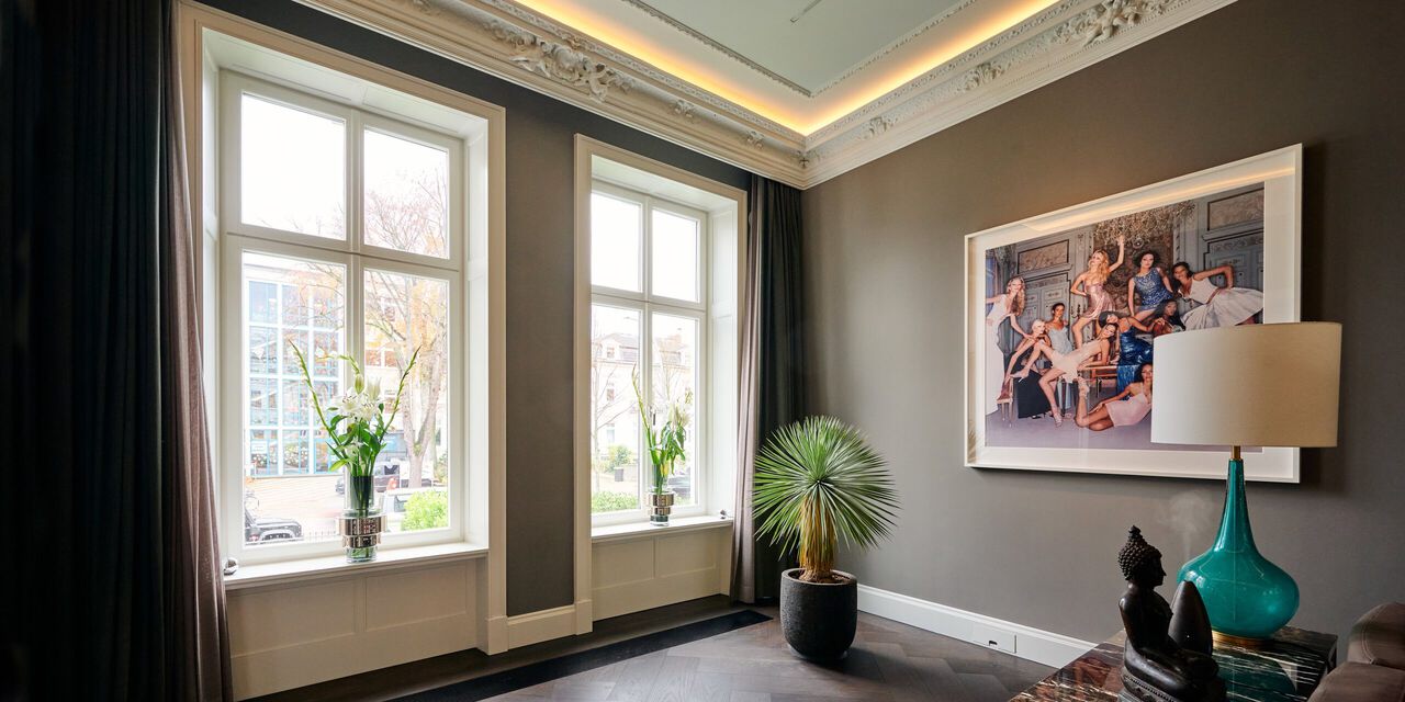 Helles Wohnzimmer mit gro&szlig;en Fenstern, Pflanzen, Kunstwerk an der Wand und eleganter Beleuchtung.