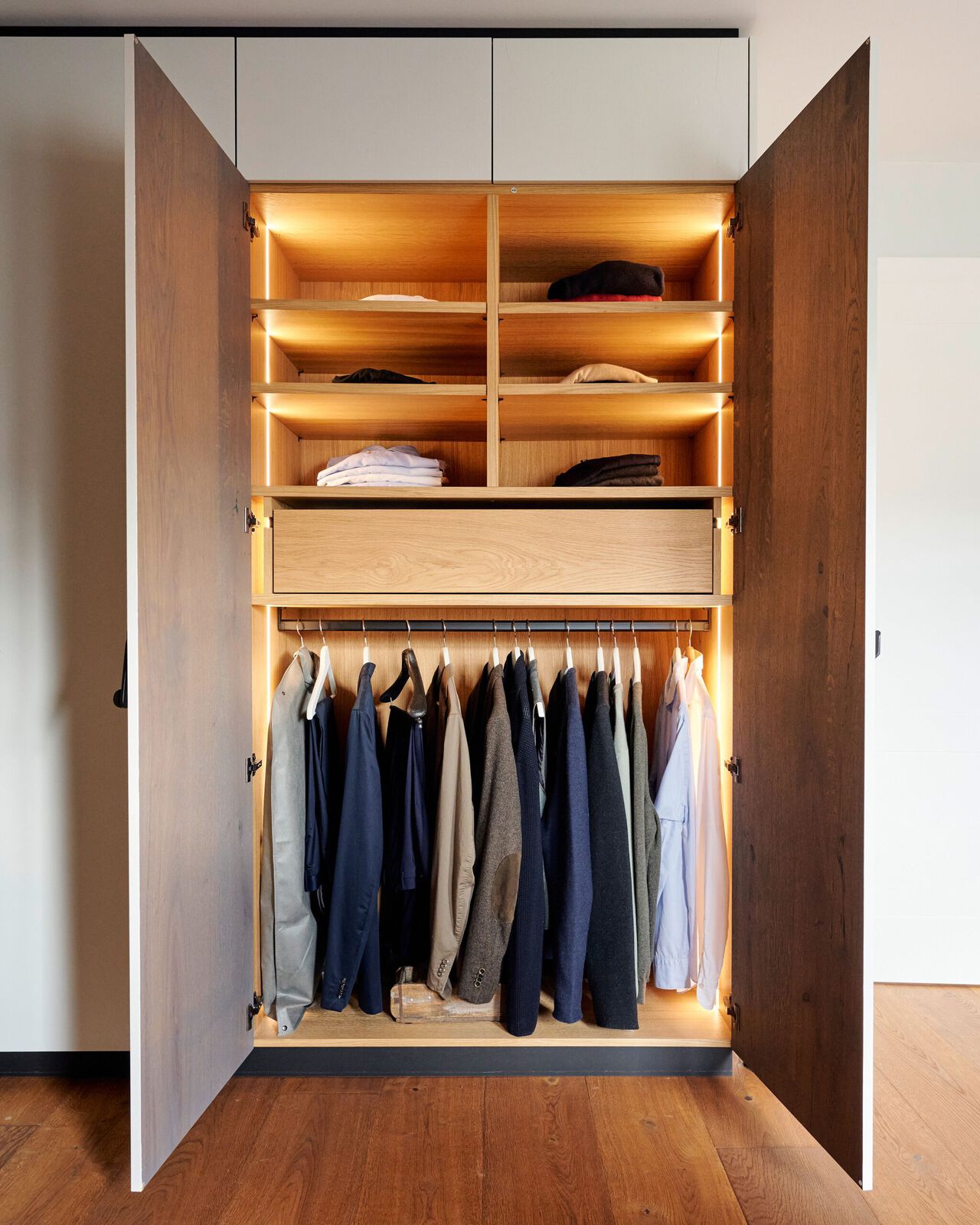 Offener Kleiderschrank aus Holz mit beleuchteten Regalen oben und Kleiderstange unten, gef&uuml;llt mit Kleidungsst&uuml;cken und gefalteter W&auml;sche.