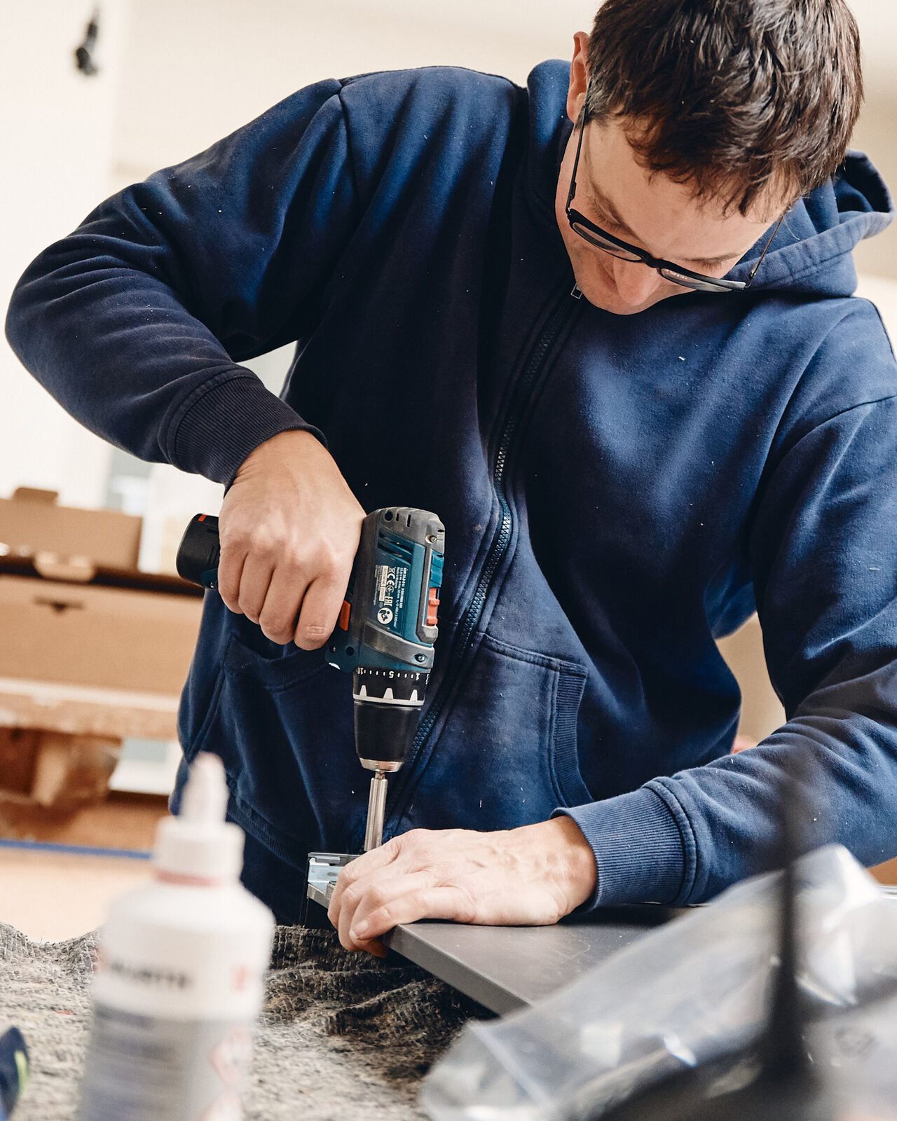 Ein Handwerker in dunklem Hemd bohrt mit einer Elektrobohrmaschine in ein Holzwerkst&uuml;ck.